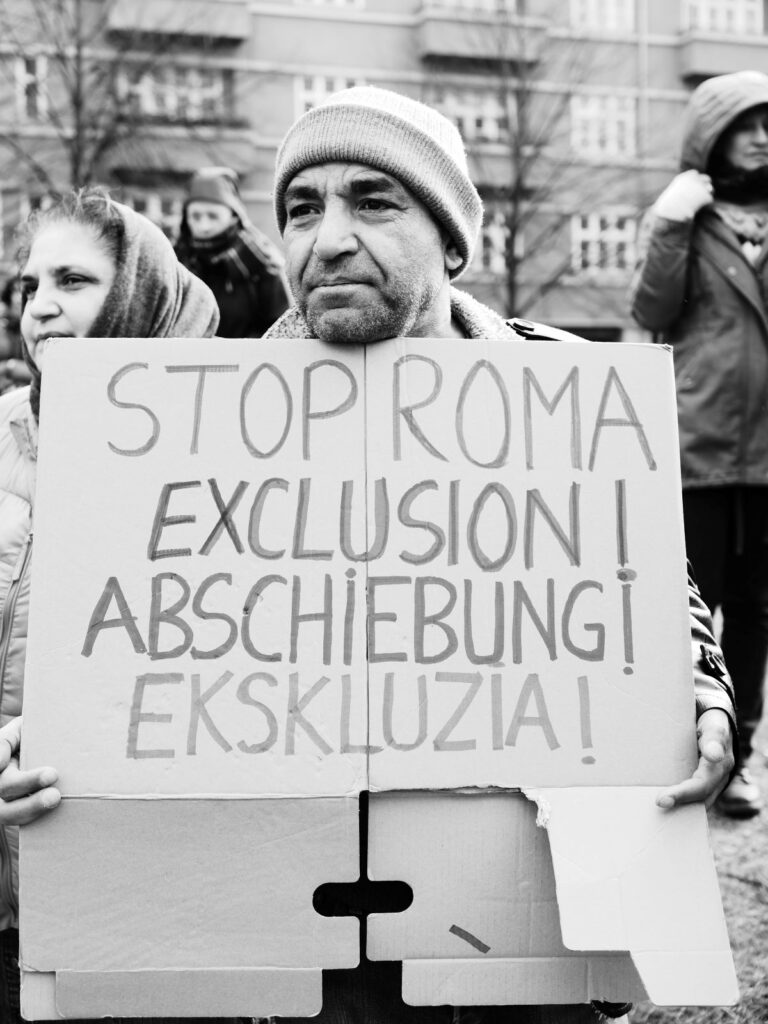 Foto vom Aktionstag zur Situation von Roma* aus der Republik Moldau. Zu sehen ist eine Person. Sie hält ein Demoschild. Auf dem steht: "Stop Roma" und folgend in den Sprachen Deutsch, Romanes und Englisch das Wort: "Abschiebung". Im Hintergrund Publikum.