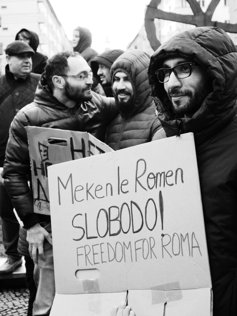 Foto vom Aktionstag zur Situation von Roma* aus der Republik Moldau. Im Hintergrund ist Publikum zu sehen. Im Vordergrund steht eine Person mit einem Demoschild, auf welchem in Romanes und Englisch steht: "Freiheit für Roma"
