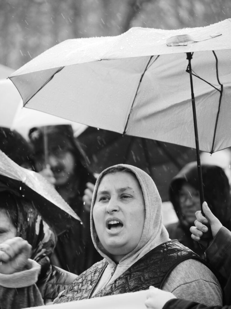 Foto von der Berlin Demonstration gegen die Abschiebung von Roma* nach Moldau. Schwarz-weiß Porträtaufnahme einer demonstrierenden weiblich gelesenen Person mit Schirm und empor gestreckter Faust.