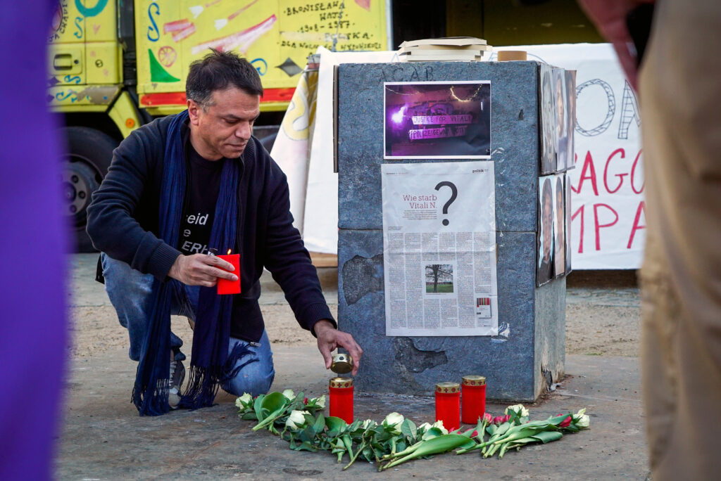 Foto von der Kundgebung für die Opfer rassistischer Polizeigewalt in Berlin. Ein Mann kniet vor einem Gedenkstein. An diesem sind Fotos und ein Zeitungsartikel angebracht. Der Artikel hat die Überschrift "Wie starb Vitali N?". 
