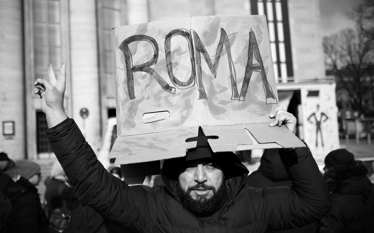 Foto vom Aktionstag zur Situation von Roma* aus der Republik Moldau. Eine Person macht das Victory-Zeichen und hält ein Demoschild mit der Aufschrift: "Roma". Im Hintergrund die Volksbühne und Publikum.