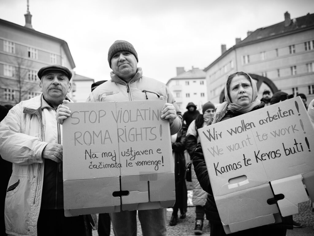 Foto vom Aktionstag zur Situation von Roma* aus der Republik Moldau. Zu sehen sind drei Personen. Sie halten Demoschilder. Auf denen steht jeweils in den Sprachen Deutsch, Romanes und Englisch: "Schluss mit der Verletzung der Roma-Rechte!" und "Wir wollen arbeiten!"