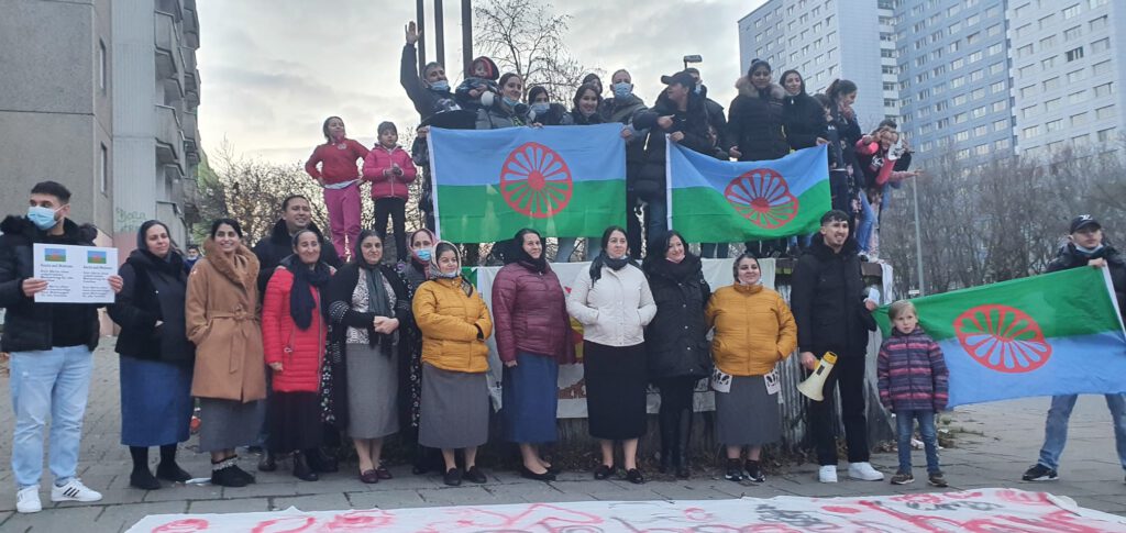 Gruppenbild Bare Berlin - Bündnis gegen Antiziganismus und für Roma*-Empowerment. Einige Personen halten eine Flagge der Roma. Diese ist Blau und Grün. In der Mitte befindet sich ein rotes Speichenrad.